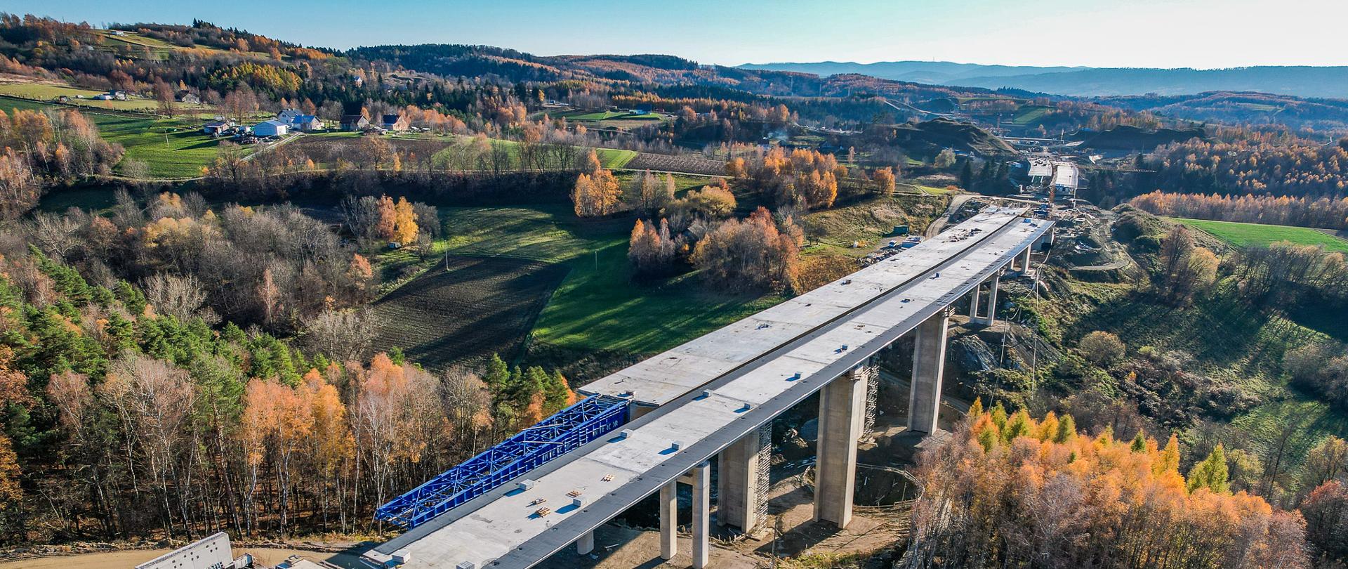 Zdjęcie z lotu ptaka przedstawia budowaną estakadę przebiegającą przez pagórkowaty, jesienny krajobraz. Betonowa konstrukcja wspiera się na wysokich filarach, a na jej początku widać niebieskie urządzenie do nasuwania segmentów. Wokół znajdują się lasy w jesiennych barwach, wykopy i teren przygotowany pod dalsze prace.