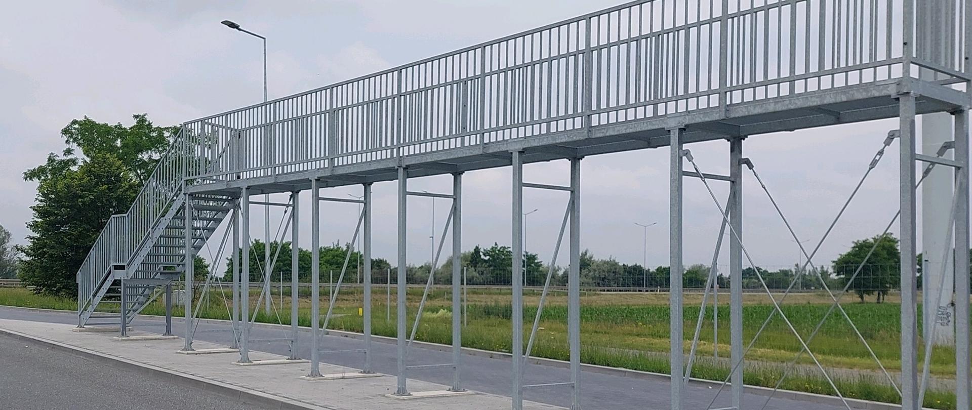 Zdjęcie przedstawia pomost wyniesiony nad ziemię. Konstrukcja obejmuje schody po lewej stronie, które umożliwiają dostęp, oraz jest wsparta na wielu pionowych i skośnych metalowych belkach. Kładka przechodzi nad asfaltową drogą, a w tle widać trawiasty teren oraz drzewa. Niebo jest pochmurne, a w pobliżu schodów znajduje się latarnia uliczna. Kładka wydaje się być zaprojektowana do bezpiecznego przechodzenia nad drogą lub do korzystania z niej przy usuwaniu śniegu z samochodów ciężarowych.