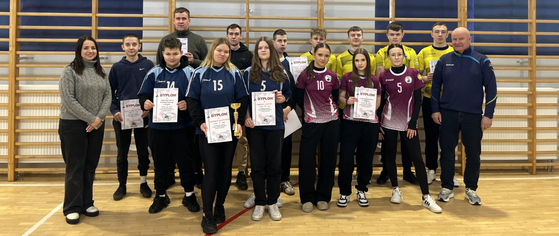 Grupowe zdjęcie uczestników i opiekunów po zawodach sportowych w sali gimnastycznej; drużyny stoją przy drabinkach i trzymają dyplomy