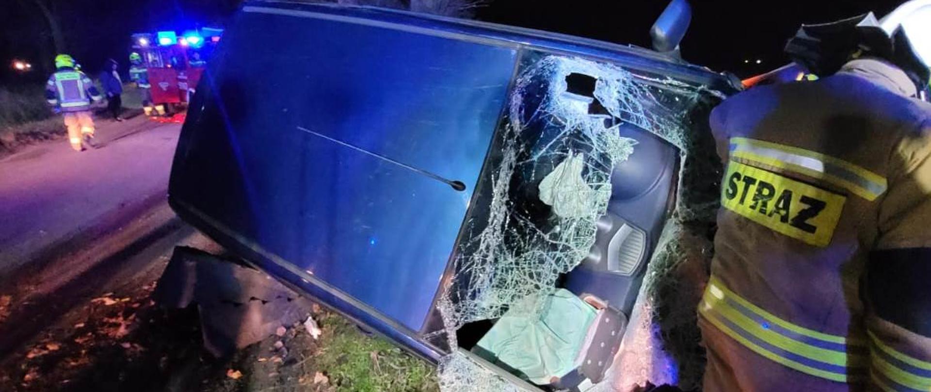 Samochód na prawym boku, uszkodzona przednia szyba, w pobliżu strażak