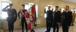Zdjęcie przedstawia trzech strażaków w mundurze wyjściowym podczas ślubowania. Ślubujący stoją w szeregu z uniesionymi prawymi dłońmi – wypowiadają sowa przysięgi. Po lewo poczet sztandarowy Komendy Powiatowej. Sztandar pochylony przed strażakami na moment ślubowania. Za ślubującymi strażacy KP PSP w Brodnicy.