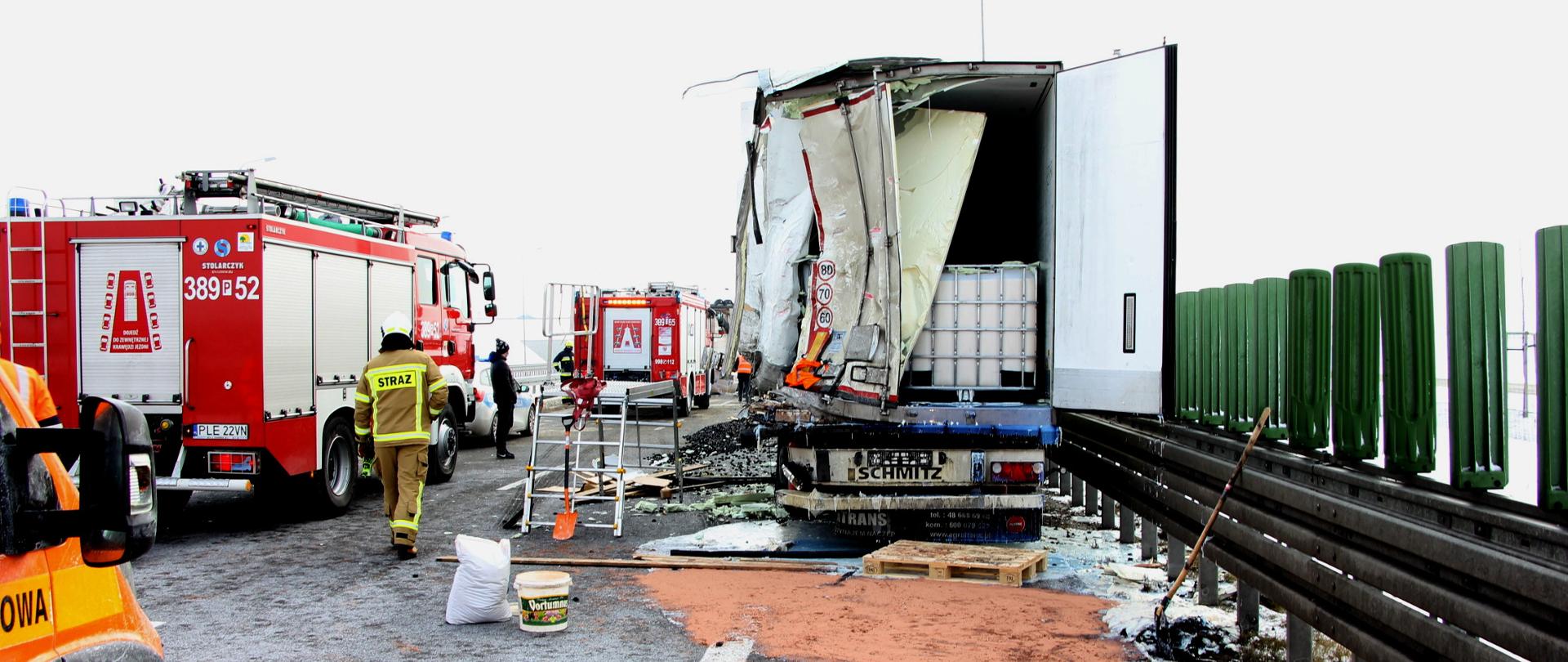 Uszkodzony samochód ciężarowy w tle drugi uszkodzony samochód ciężarowy oraz samochody pożarnicze.