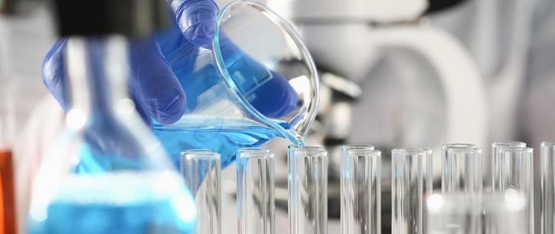 Osoba w rękawiczkach laboratoryjnych przelewa niebieski roztwór z kolby do probówek ustawionych w rzędzie; w tle widoczne szkło laboratoryjne i wyposażenie laboratorium.