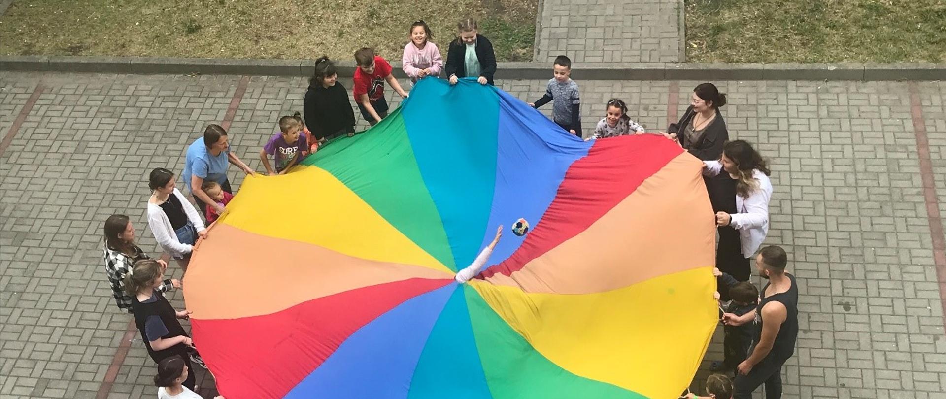 Widok z góry. Grupa dzieci stoi i trzyma wielką, kolorowa okrągłą chustę. Ze środka chusty wystaje ręka.