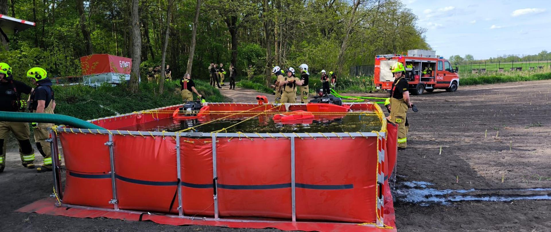 Na zdjęciu widzimy grupę strażaków podczas ćwiczeń na terenie leśnym. Na pierwszym planie dwóch strażaków w hełmach w kolorze jaskrawej zieleni pracuje wspólnie przy obsłudze pompy pożarniczej. Obaj strażacy maja na sobie mundury strażackie z krótkimi rękawami jeden z nich wskazuje coś na pompie a drugi dokładnie się temu przygląda co świadczy o koncentracji i precyzji działania.