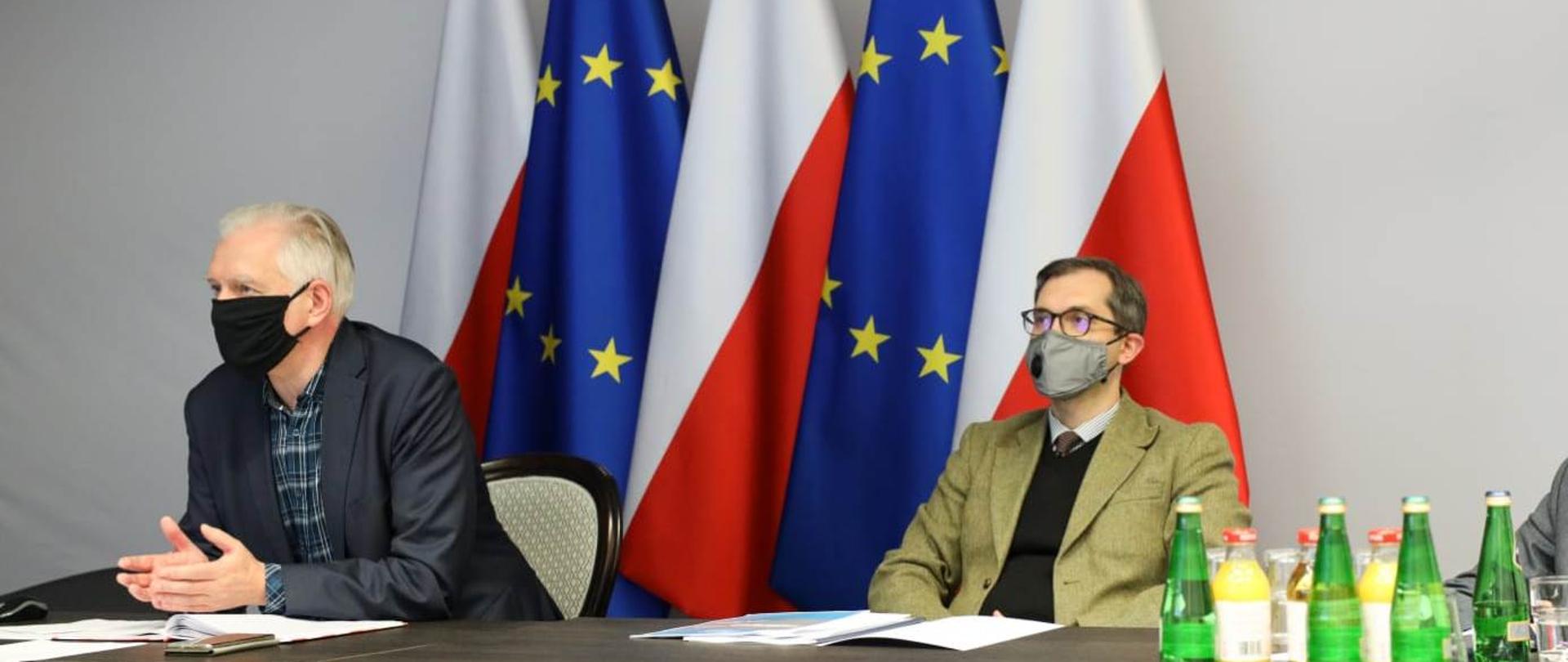 Na zdjęciu wicepremier Jarosław Gowin i wiceminister Marek Niedużak podczas wideokonferencji. Siedzą za stołem, mają maseczki na twarzach, w tle fagi Polski i UE.