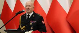 Przy mównicy strażak w mundurze galowym. Obok dekoracja z biało-czerwonych kwiatów, w tle kilka flag, stojących w szeregu, biało-czerwonych.