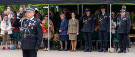 Na pierwszym planie w centrum stoi tyłem do obiektywu dowódca apelu wyprostowany na baczność, podczas gdy w tle, pod czarnym namiotem, na krzesłach siedzą i stoją zaproszeni goście honorowi w strojach wizytowych oraz mundurach. Dowódca ubrany jest w galowy mundur strażacki ze sznurami, baretkami i czarnymi skórzanymi oficerkami z wysoką cholewą. Tło wypełnia czarne zadaszenie, niska zieleń ozdobna wokół wybrukowanego placu oraz ściana budynku, bez umieszczonych napisów. Pogoda jest stabilna i pochmurna, z łagodnym światłem. Ujęcie to emanuje rygorem wojskowo-strażackim, powagą, dyscypliną oraz uroczystym szacunkiem dla zgromadzonych gości.