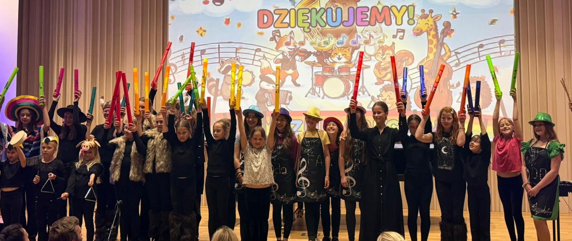 Na scenie stoi grupa dzieci podczas występu muzycznego. Ubrane są w jednakowe czarne stroje i trzymają bum-bum-rurki. Dzieci śpiewają, stojąc w rzędzie przed publicznością. W tle wyświetlona jest kolorowa grafika z napisem dziękujemy. Na widowni widoczne są sylwetki oglądających. Atmosfera wydarzenia jest radosna i koncertowa.