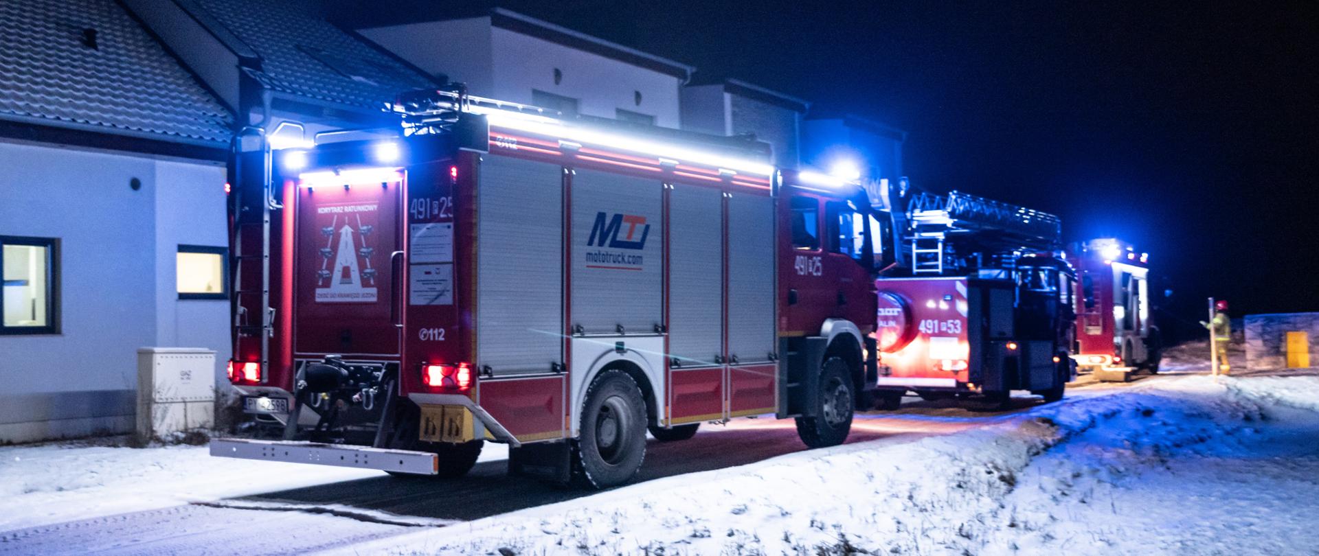 Przed kompleksem restauracyjno - hotelowym stoją wozy strazackie. Pojazdy mają włączone światła zewnętrzne oraz lampy błyskowe. W oddali widoczny strażak który ocenia sytuację. Zimowa aura w tle.