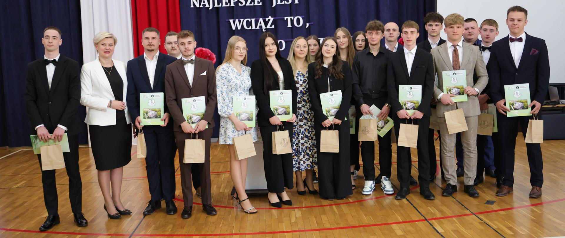 Uczniowie i nauczyciele pozują do wspólnego zdjęcia podczas szkolnej uroczystości, trzymając pamiątkowe materiały.