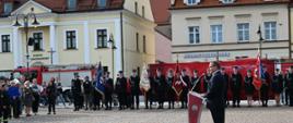 Na pierwszym planie, z prawej strony, stoi mężczyzna w ciemnym garniturze, który przemawia do zgromadzonych przy nowoczesnej, czerwonej mównicy z logo Państwowej Straży Pożarnej. W tle, naprzeciwko mówcy, ustawione są poczty sztandarowe oraz rzędy strażaków w czarnych mundurach galowych, stojących na baczność przed dużą czerwoną ścianką promocyjną. Po lewej stronie placu widoczna jest grupa mieszkańców obserwujących wydarzenie, a za nimi zaparkowany wóz strażacki oraz zabytkowe kamienice.