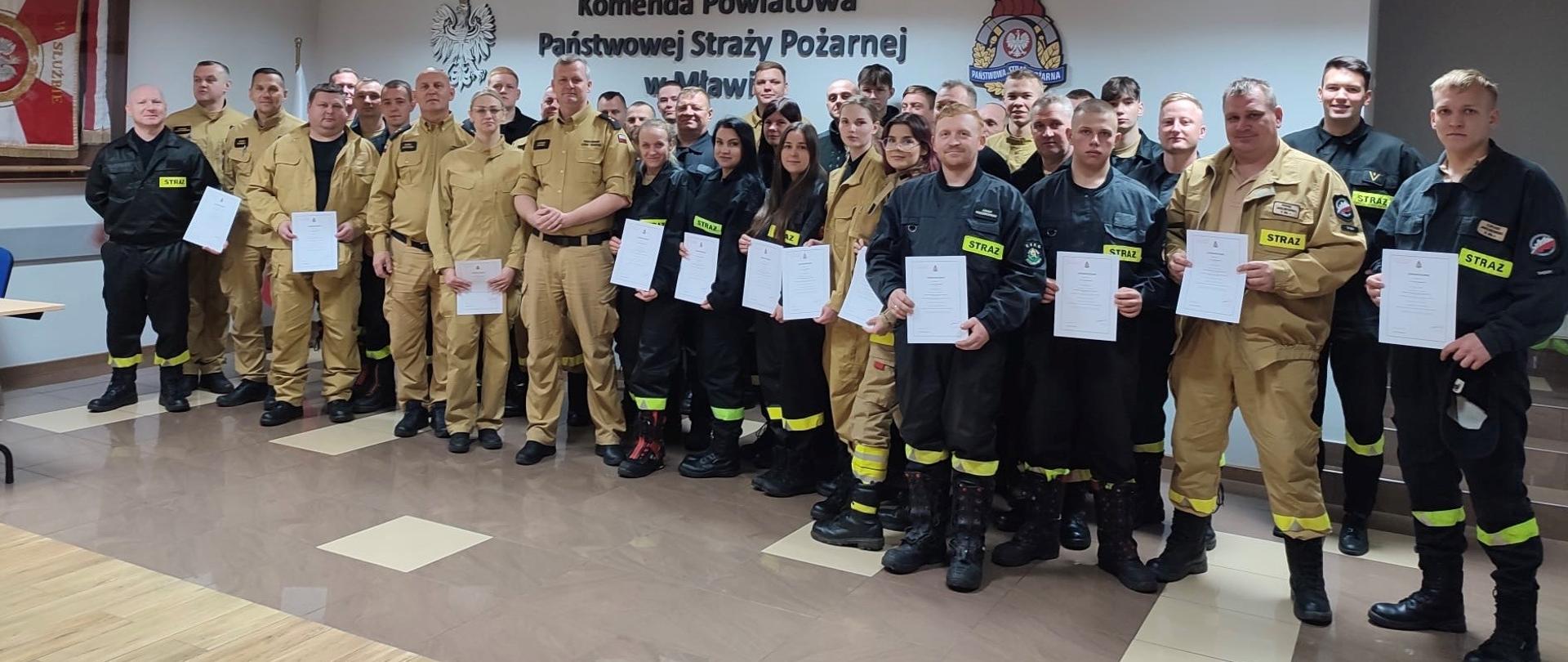 Na zdjęciu widać grupę strażaków pozujących do wspólnego zdjęcia w pomieszczeniu komendy Państwowej Straży Pożarnej w Mławie. Część osób ubrana jest w czarne, a część w beżowe mundury strażackie. Wiele z nich trzyma w rękach certyfikaty lub dyplomy. W tle widoczny jest napis „Komenda Powiatowa Państwowej Straży Pożarnej w Mławie” oraz godła i symbole straży pożarnej. Zdjęcie ma charakter pamiątkowy, prawdopodobnie wykonane po zakończeniu szkolenia lub wręczeniu zaświadczeń.