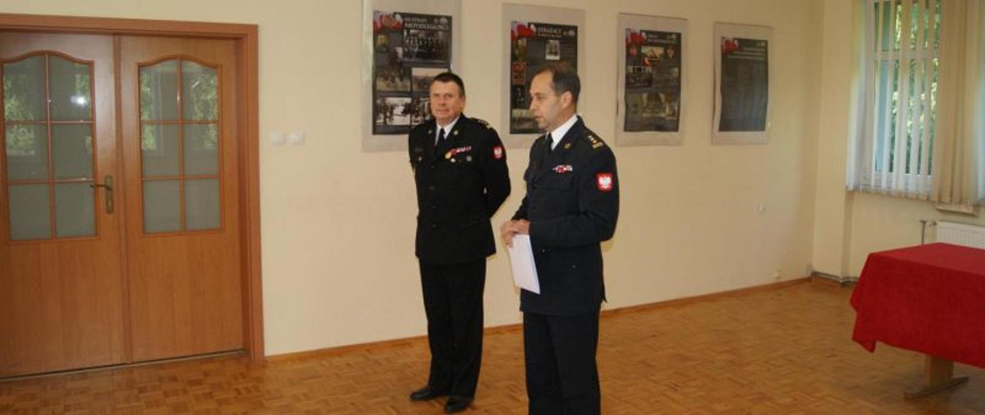 Centralny punkt zdjęcia stanowi dwóch strażaków. Komendant przyjmujący obowiązki od swojego przełożonego i przełożony. Wydarzenie ma miejsce wewnątrz budynku, na świetlicy. Obydwaj komendanci w galowych, wyjściowych mundurach.