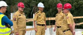 Małopolski Komendant Wojewódzki PSP wraz z zastępcami oraz Komendantem Powiatowym PSP w Wadowicach w mundurach służbowych stoją na terenie budowy w towarzystwie pracownika budowy. Widoczne zbrojenia i drewniane barierki zabezpieczające. W tle budowane filary z widocznym zbrojeniem 