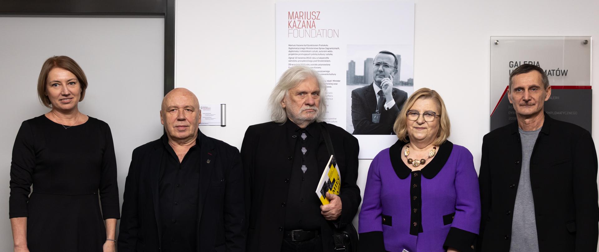Na zdjęciu jest 5 osób - dwie kobiety i trzech mężczyzn. Wszyscy patrzą w obiektyw. Mają na sobie ciemne ubrania. Za nimi jest tabliczka z napisem "Galeria u Dyplomatów" oraz plakat z napisem "Mariusz Kazana Foundation". Mężczyzna w środku trzyma żółtą broszurę. 