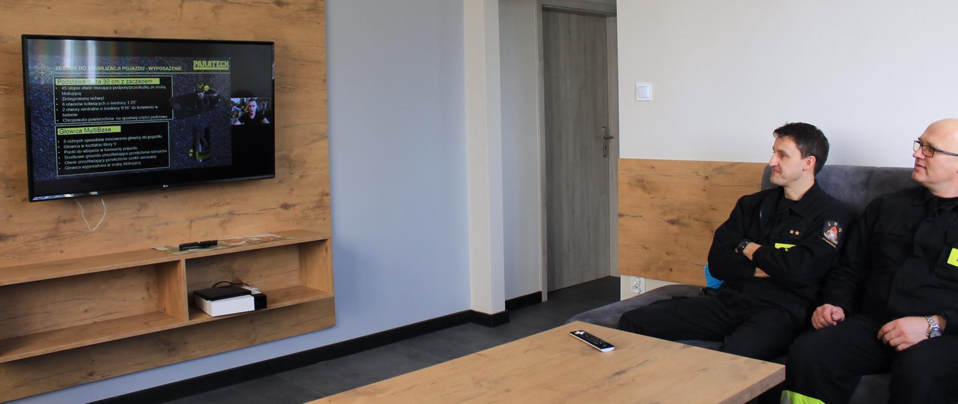 Strażacy uczestniczą w wideo-szkoleniu. Po lewej stronie zdjęcia na ścianie jest telewizor na którym wyświetlane są tabele. Naprzeciwko telewizora na kanapie siedzą strażacy. 