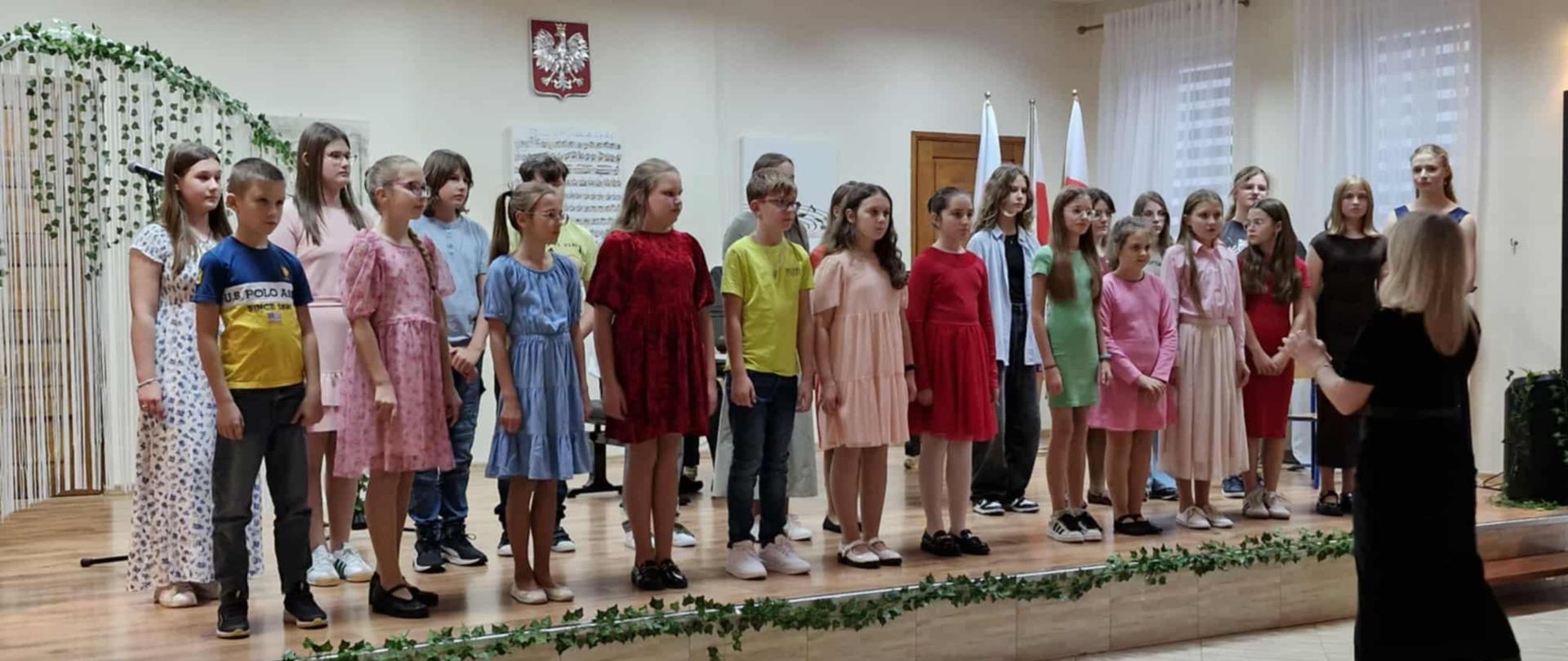 Chór szkolny podczas występu. Grupa dzieci w strojach galowych śpiewa, stojąc na podwyższeniu na scenie. Przed nimi stoi ubrana na czarno młoda dyrygentka.