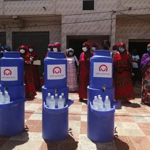 na podłodze stoją trzy pojemniki na wodę, a w tle stoją kobiety ubrane w kolorowe ubrania