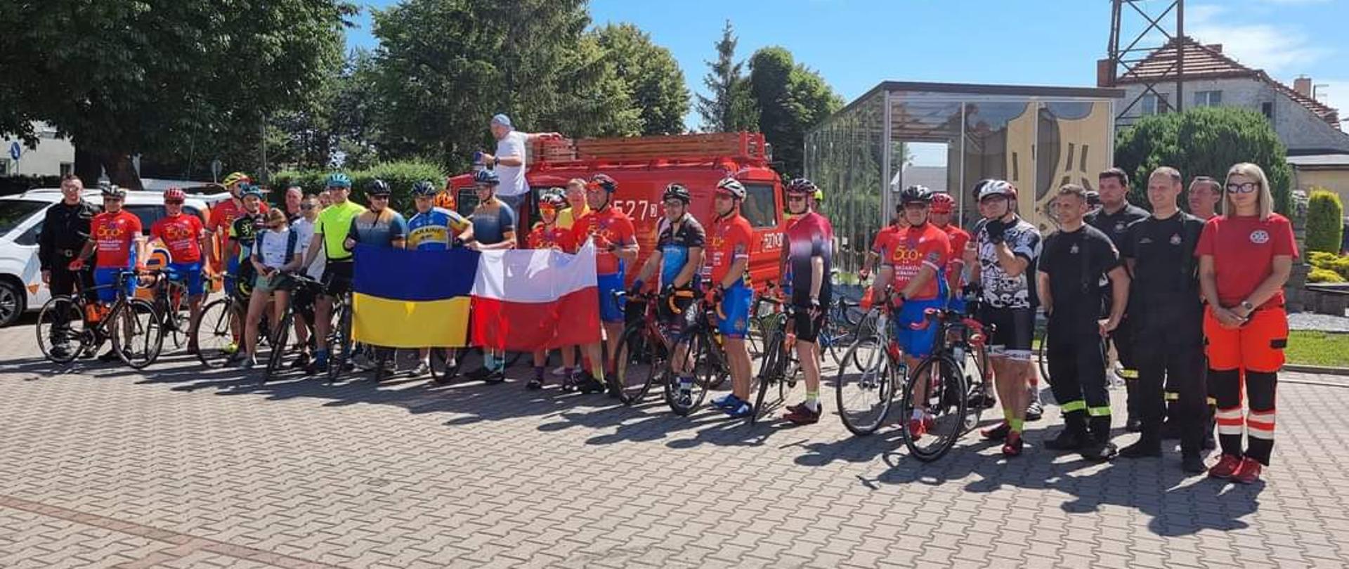 na zdjęciu widać grupę kolarzy - strażaków na placu Komendy PSP w Oławie. Trzymają flagę Polski i Ukrainy.