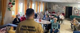 Strażak omawia zagrożenia występujące w prywatnych domach mieszkalnych przy użyciu prezentacji multimedialnej. Seniorzy siedzą i słuchają prelegenta.