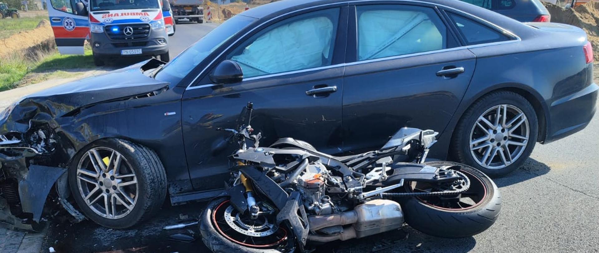 Zdjęcie przedstawia wypadek z udziałem samochodu osobowego z motocyklem. Motocykl leży przed autem. Samochód ma uszkodzony przód. Widać plamę płynów eksploatacyjnych. Z tyłu widać karetkę pogotowia. 