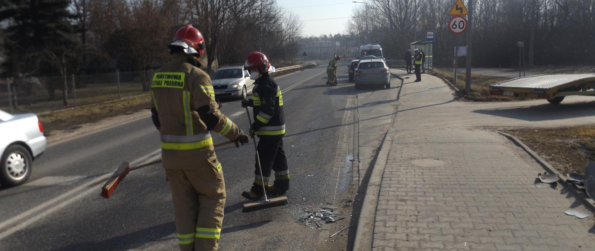 Zdjęcie przedstawia strażaków porządkujących teren akcji po kolizji. W tle widać lawetę oraz policjanta i karetkę Pogotowia Ratunkowego. 