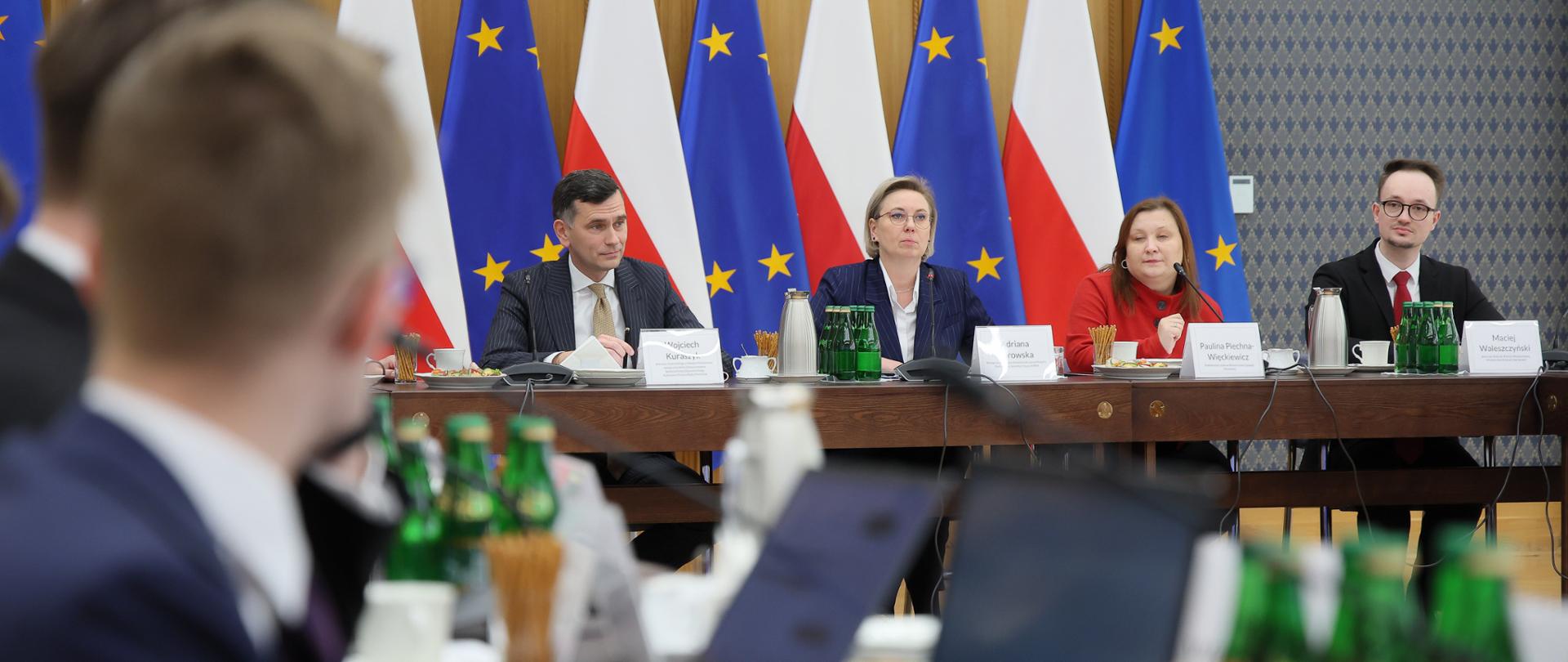 Zdjęcie sytuacyjne. Na zdjęciu uczestnicy posiedzenia Rady Dialogu z Młodym Pokoleniem obradują przy stole konferencyjnym. Na dalszym planie, widoczni są od lewej: Wojciech Kuraszyk, Zastępca Dyrektora w Departamencie Społeczeństwa Obywatelskiego w KPRM; Adriana Porowska, Sekretarz Stanu w KPRM i Wiceprzewodnicząca Komitetu do spraw Pożytku Publicznego, Paulina Piechna-Więckiewicz, Podsekretarz Stanu w Ministerstwie Edukacji Narodowej oraz Maciej Waleszczyński, Sekretarz Rady do spraw Polityki Młodzieżowej, Ministerstwo Edukacji Narodowej. W tle widoczne są flagi Polski i Unii Europejskiej.