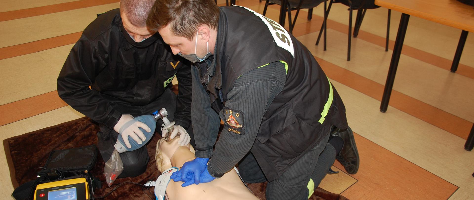 Na pierwszym zdjęciu strażacy-ratownicy wykonują symulację resuscytacji krążeniowo-oddechową u osoby dorosłej z użyciem defibrylatora zautomatyzowanego AED. Jeden z ratowników wykonuje 30 uciśnięć klatki piersiowej, natomiast drugi wykonuje 2 oddechy ratownicze za pomocą worka samorozprężalnego.