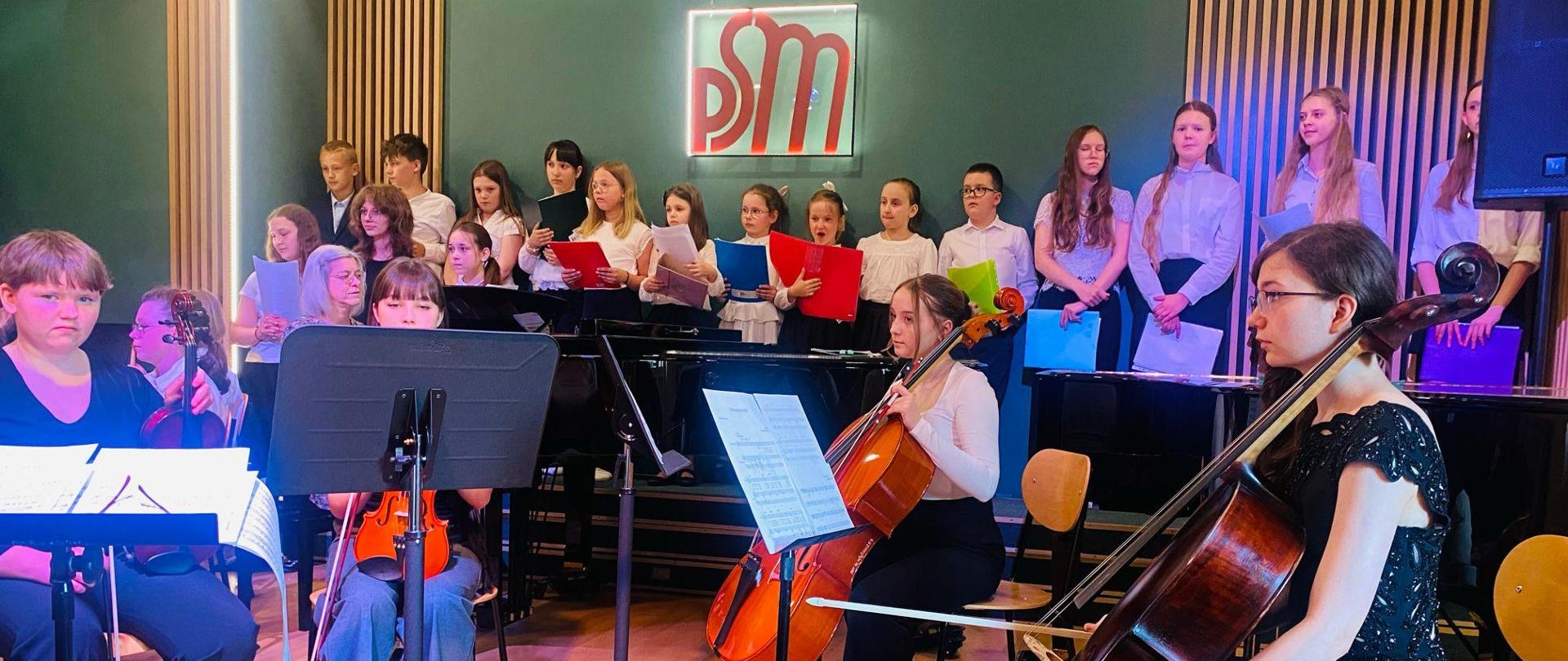 Na zdjęciu prezentuje się orkiestra szkolna oraz chór przed każdym uczniem stoi pulpit a uczniowie trzymają w rękach wiolonczele i skrzypce a chór tuż za orkiestrą trzyma w rękach kolorowe teczki w tle widać zieloną ścianę z logo szkoły