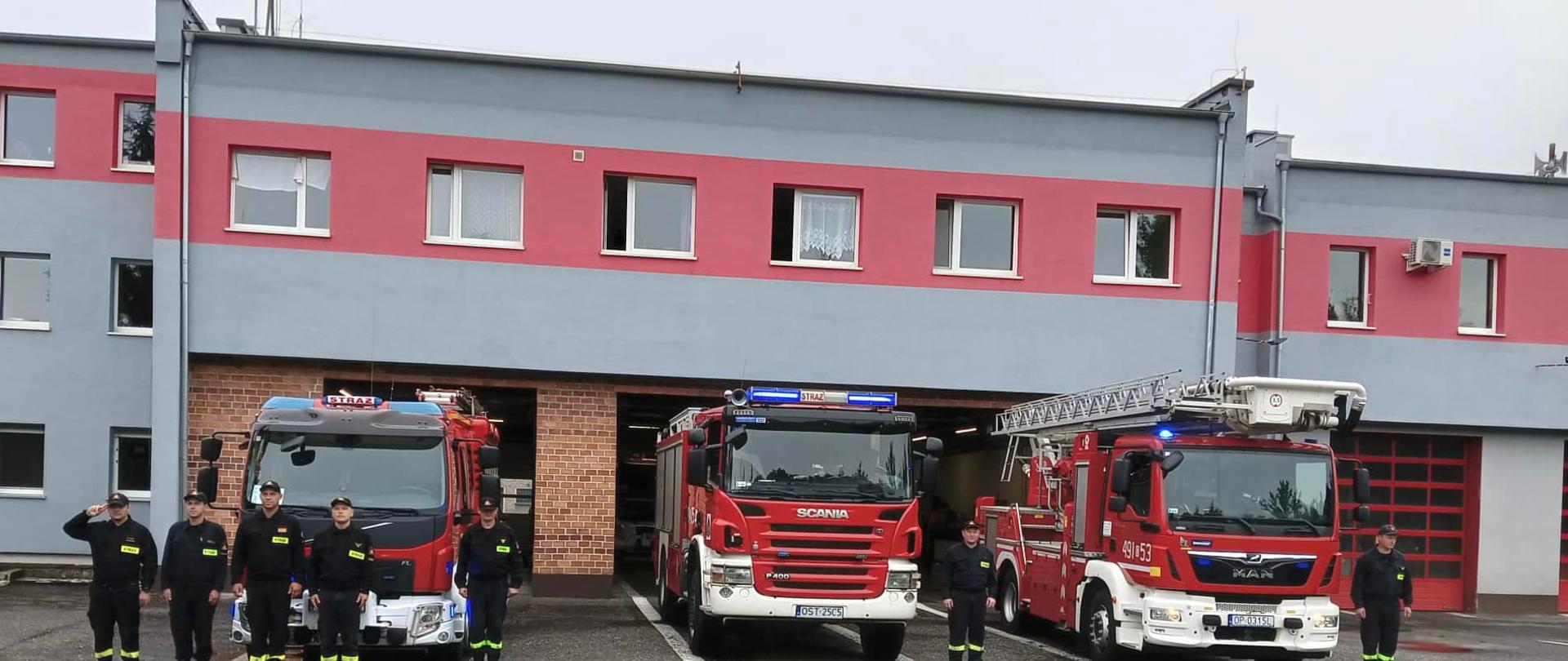 Na zdjęciu znajdują się strażacy ustawieni przed pojazdami pożarniczymi na tle budynku KP PSP w Strzelcach Opolskich
