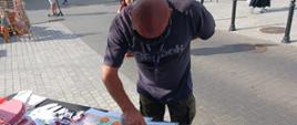 Na zdjęciu widoczny jest mężczyzna z założonymi narkogoglami stojący przy stoisku z ulotkami.