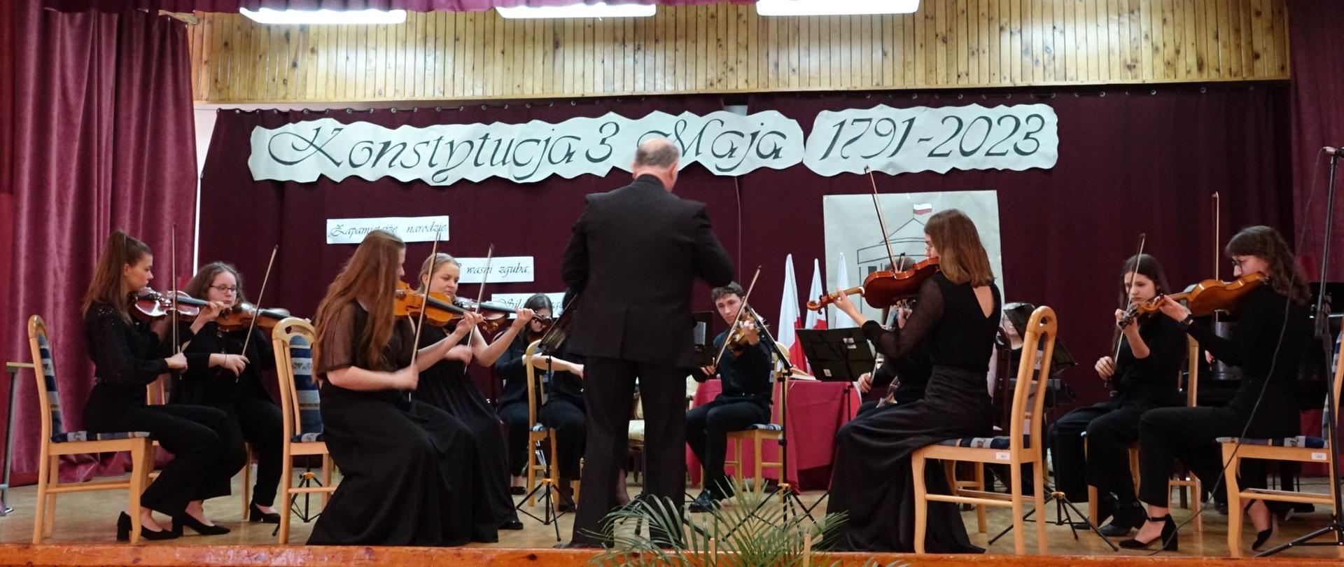 Zdjęcie. Orkiestra, którą prowadzi dyrygent, występuje na scenie. Na pierwszym planie kilkoro uczniów grających na skrzypcach. W tle napis: Konstytucja 3 Maja 1791-2023.