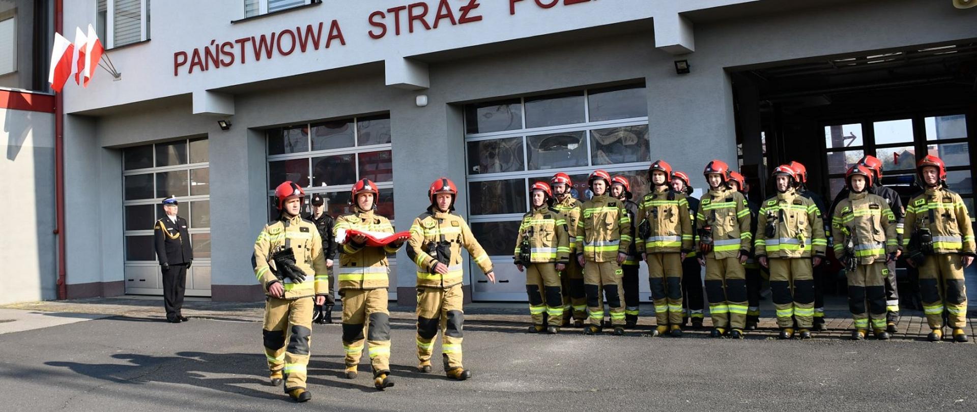 Na pierwszym planie zdjęciu widzimy trzech strażaków, którzy maszerują z flagą. Za nimi stoją strażacy w dwuszeregu oraz dowódca i komendant. W tle widzimy budynek komendy.