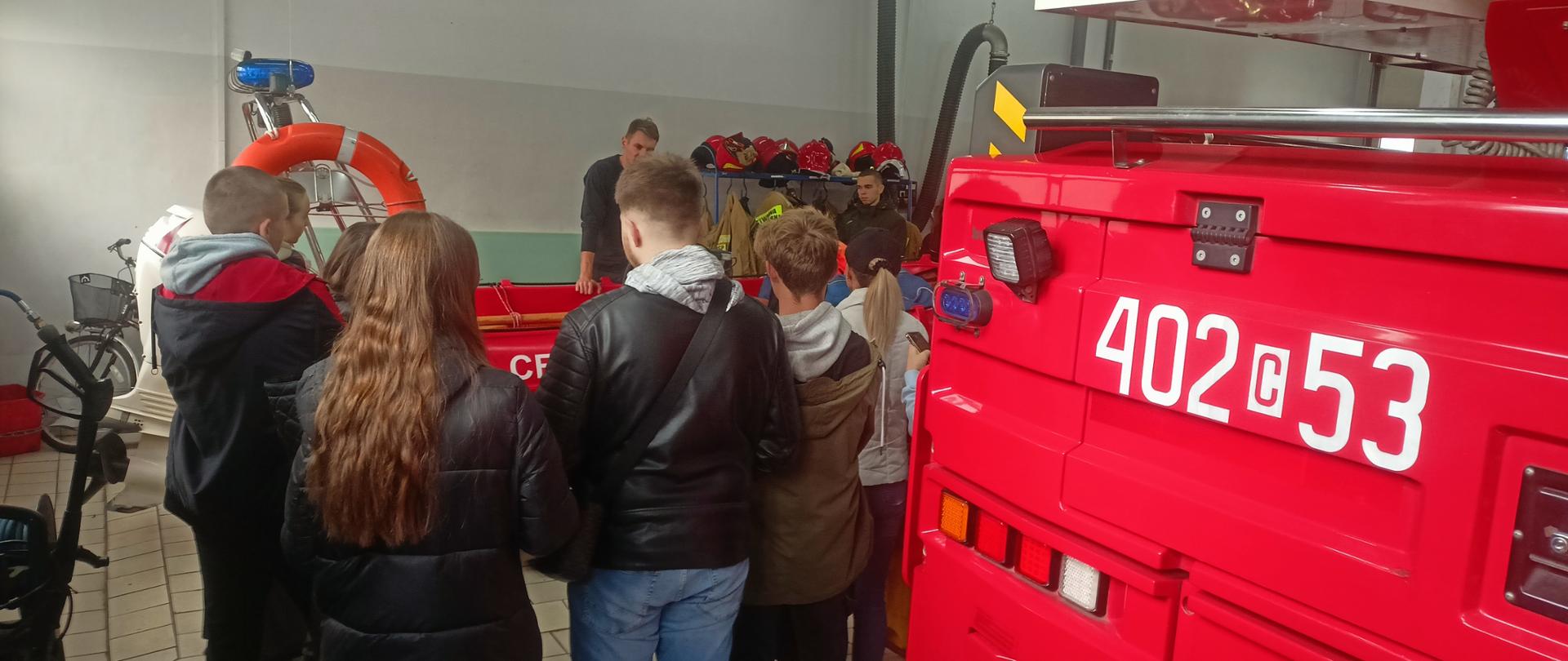 Zdjęcie przedstawia grupę młodzieży, która słucha strażaka omawiającego sprzęt będący na wyposażeniu samochodu gaśniczego GBA 2,5/16.