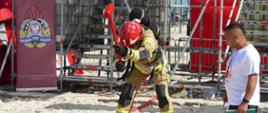 W środku zdjęcia widać strażaka w ubraniu specjalnym w kolorze pisakowym. Strażak bierze zamach młotem. Stanowisko znajduje się na plaży. Po lewej stronie reklama PSP. W tle widać rusztowania. Po prawej stronie sędzia zawodów w koszulce koloru białego i zielonych spodenkach.