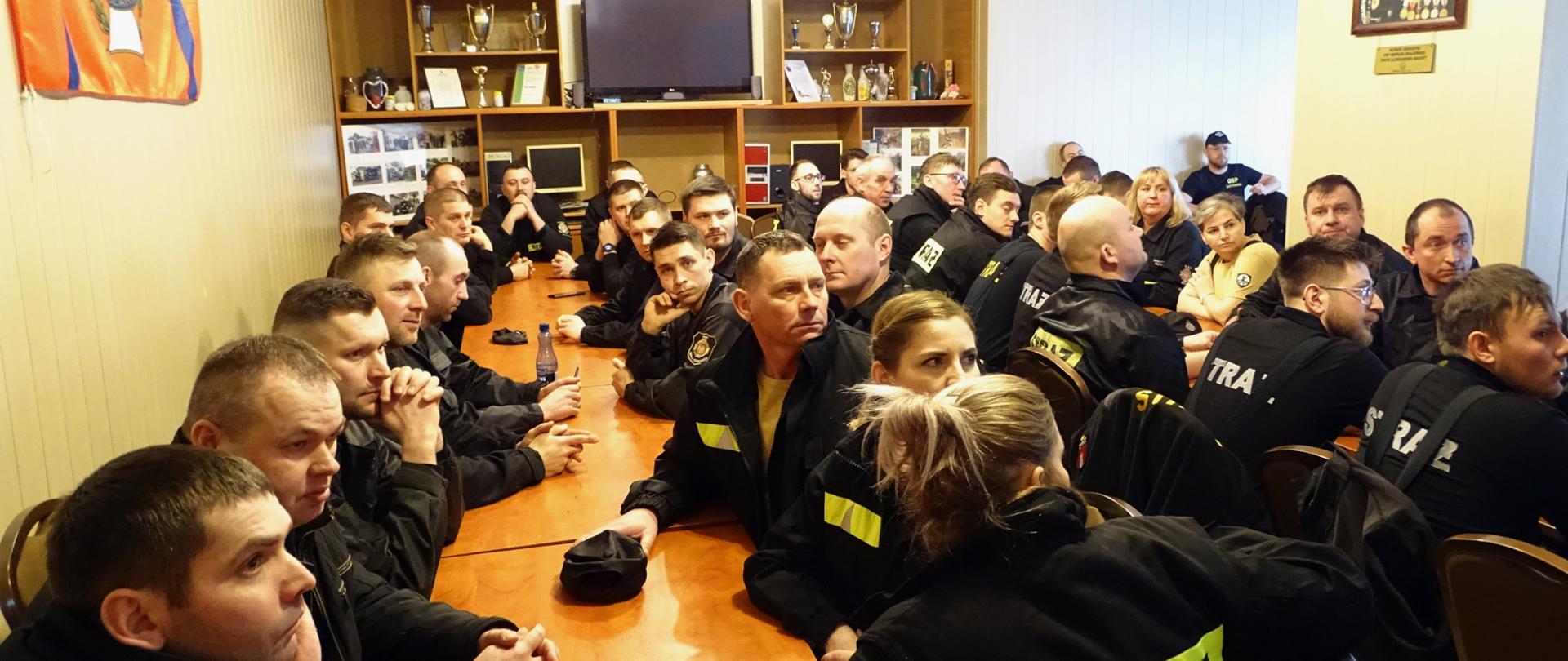 Zdjęcie wykonano w budynku siedziby Ochotniczej Straży Pożarnej w pomieszczeniu świetlicy. Strażacy Ochotniczej straży pożarnej ubrani w mundury koszarowe siedzą przy stołach czekając na egzamin.