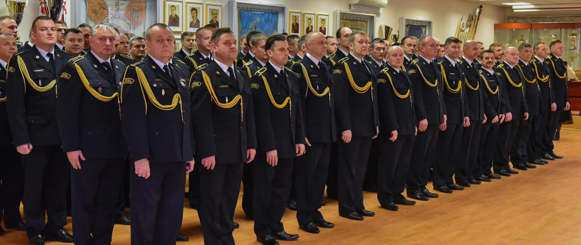 Zdjęcie przedstawia funkcjonariuszy Państwowej Straży Pożarnej stojących na zbiórce w umundurowaniach wyjściowych bez nakrycia głowy.