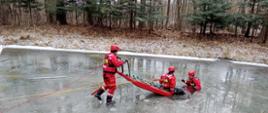 Widok na zamarznięty zbiornik wodny. Dwóch ratowników ćwiczy ratowanie tonącego w przerębli, z użyciem sań lodowych.