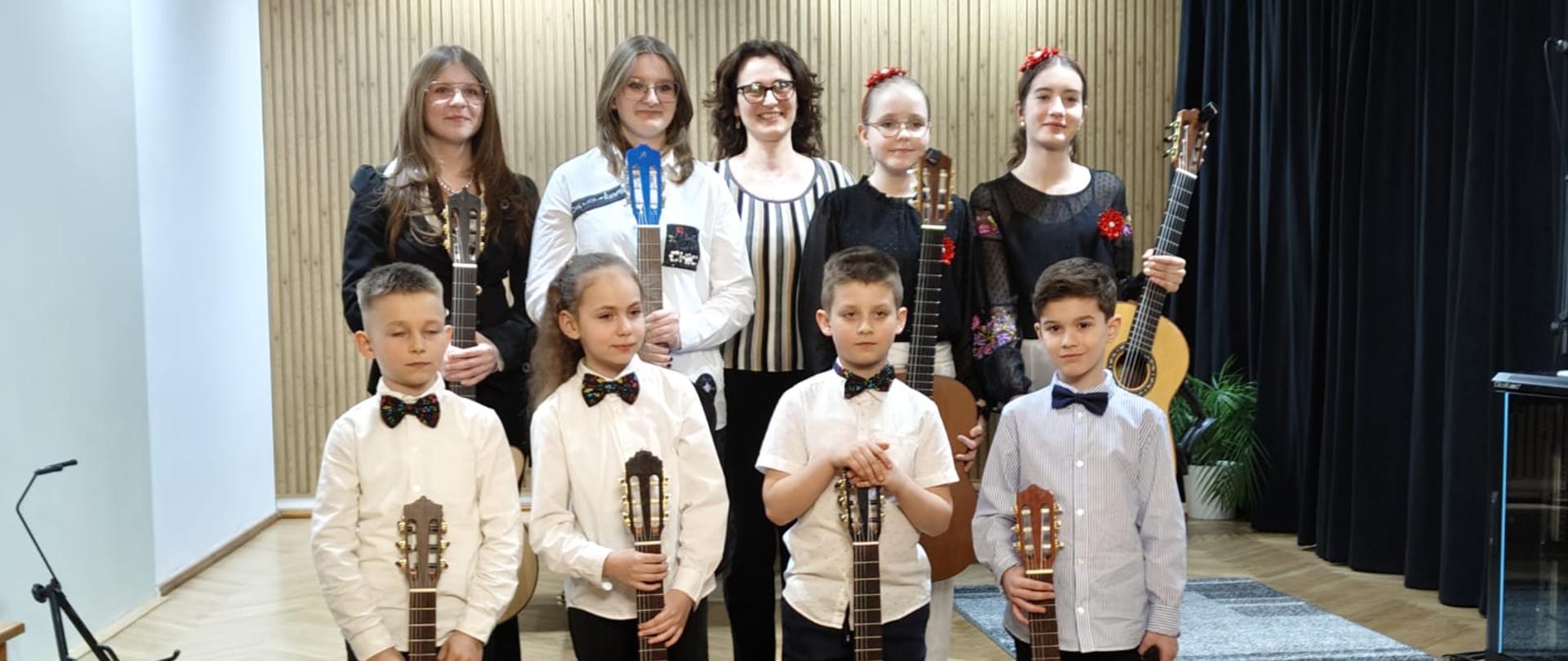 Galeria 14 zdjęć z wiosennego koncertu gitarowego uczniów klasy gitary pani Agnieszki Kozakowskiej, który odbył się 28 marca 2025 w auli PSM w Kłodzku. Uczniowie grają na gitarze na sali koncertowej.