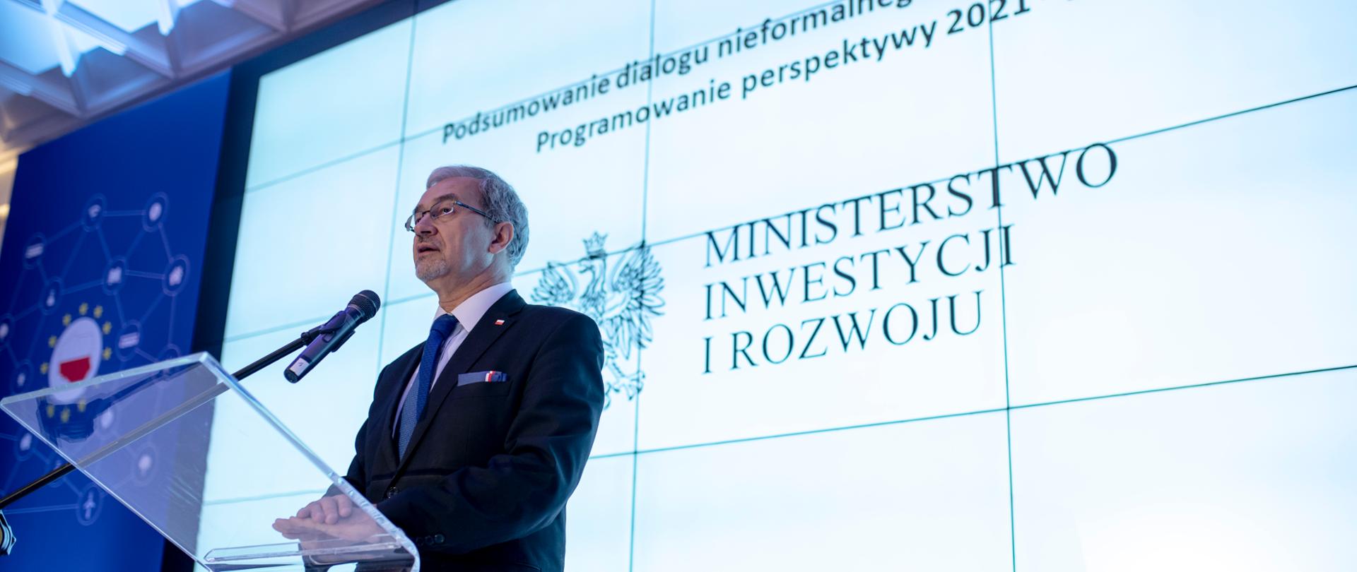 Minister stoi przy mównicy na ścianie wyświetlany jest napis "podsumowanie dialogu nieformalnego z Komisją Europejską. Programowanie perspektywy 2021-2027"