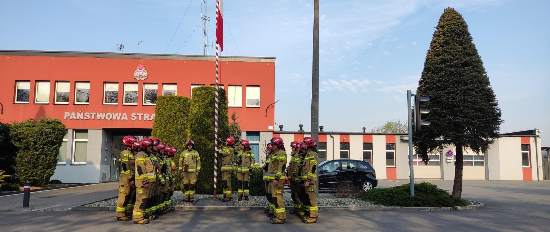 Zdjęcie przedstawia strażaków wieszających flagę państwową na tle komendy