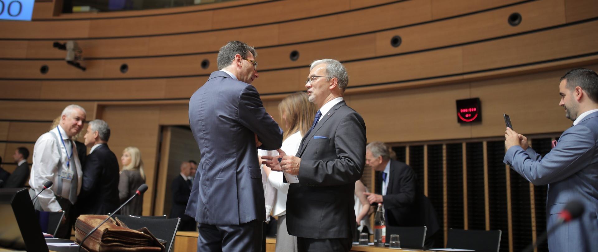 W centralnym punkcie zdjęcie stoją od lewej przedstawiciel Komisji Europejskiej Marc Lemaitre i minister Kwieciński. Rozmawiają ze sobą. Zdjęcie zrobione w sali posiedzeń Rady UE. Na sali obecni tez inni ludzie