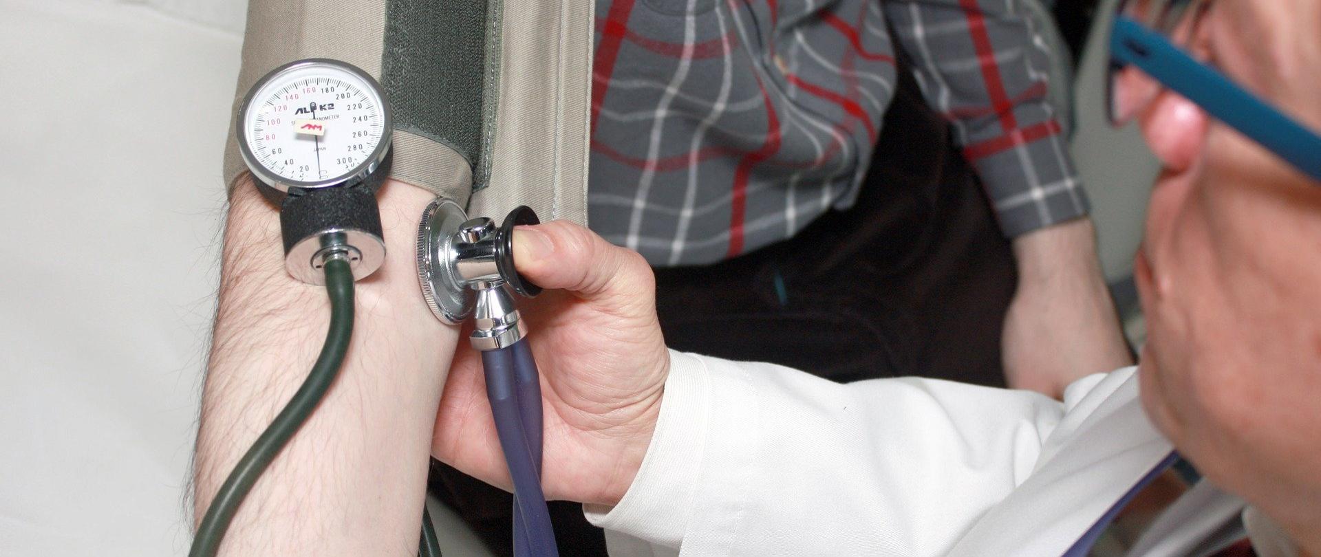 Na zdjęciu przedramię a na nim ciśnieniomierz. Po stronie prawej kadru z pozycji odgórnej widać lekarza, wykonującego pomiar ciśnienia tętniczego krwi. 