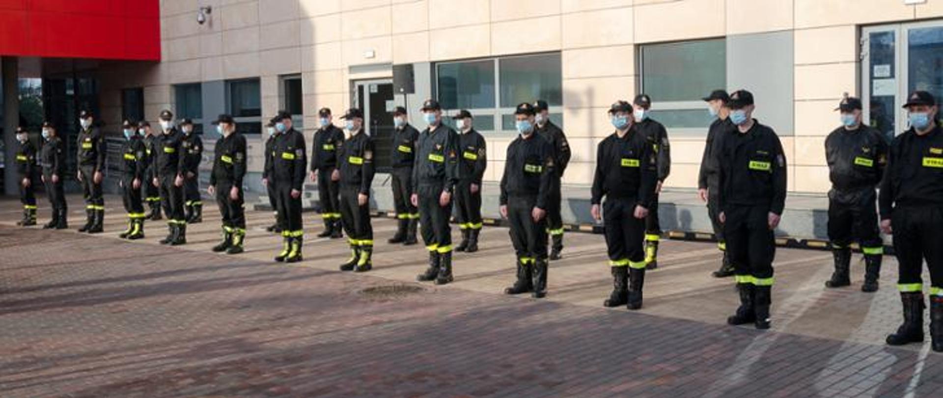 Zdjęcie przedstawia grupę strażaków stojących w dwuszeregu na tle budynku KW PSP w Warszawie