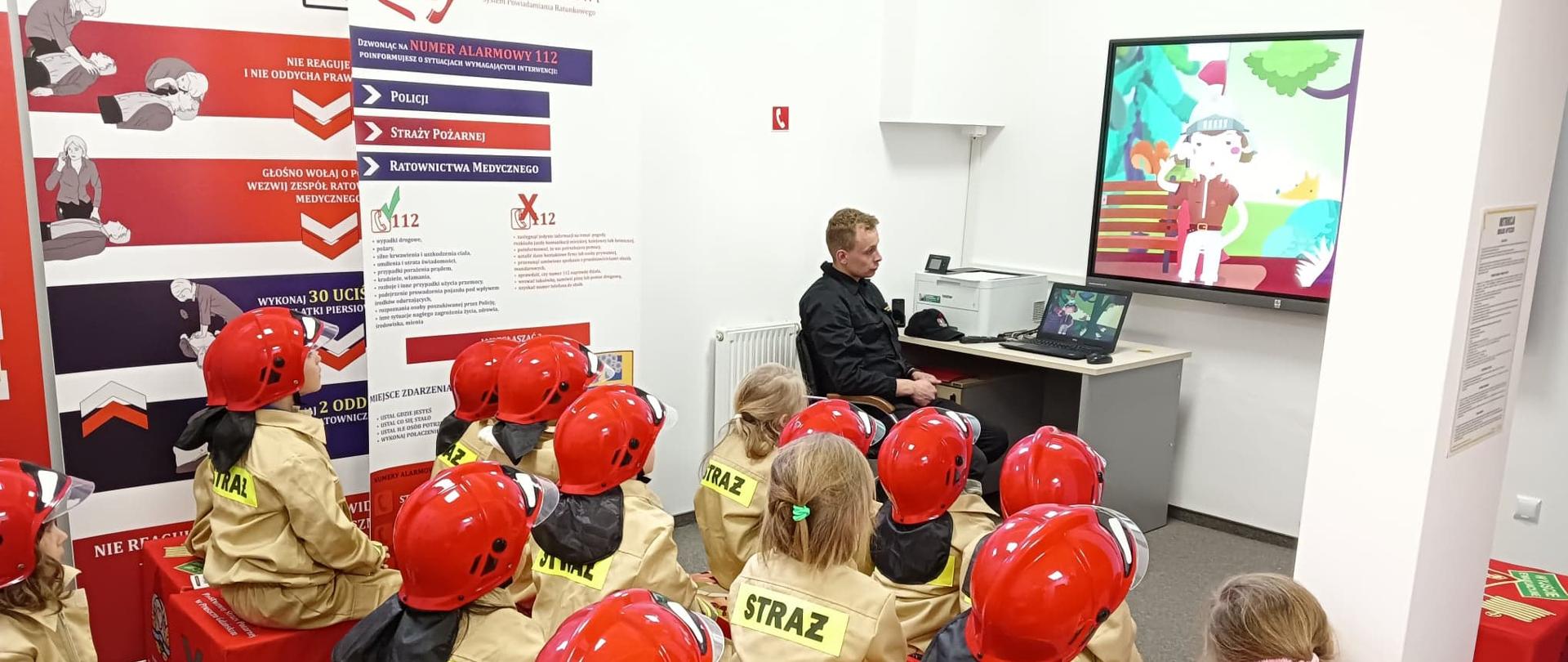 Na zdjęciu widzimy strażaka i grupę dzieci podczas oglądania filmów edukacyjnych.