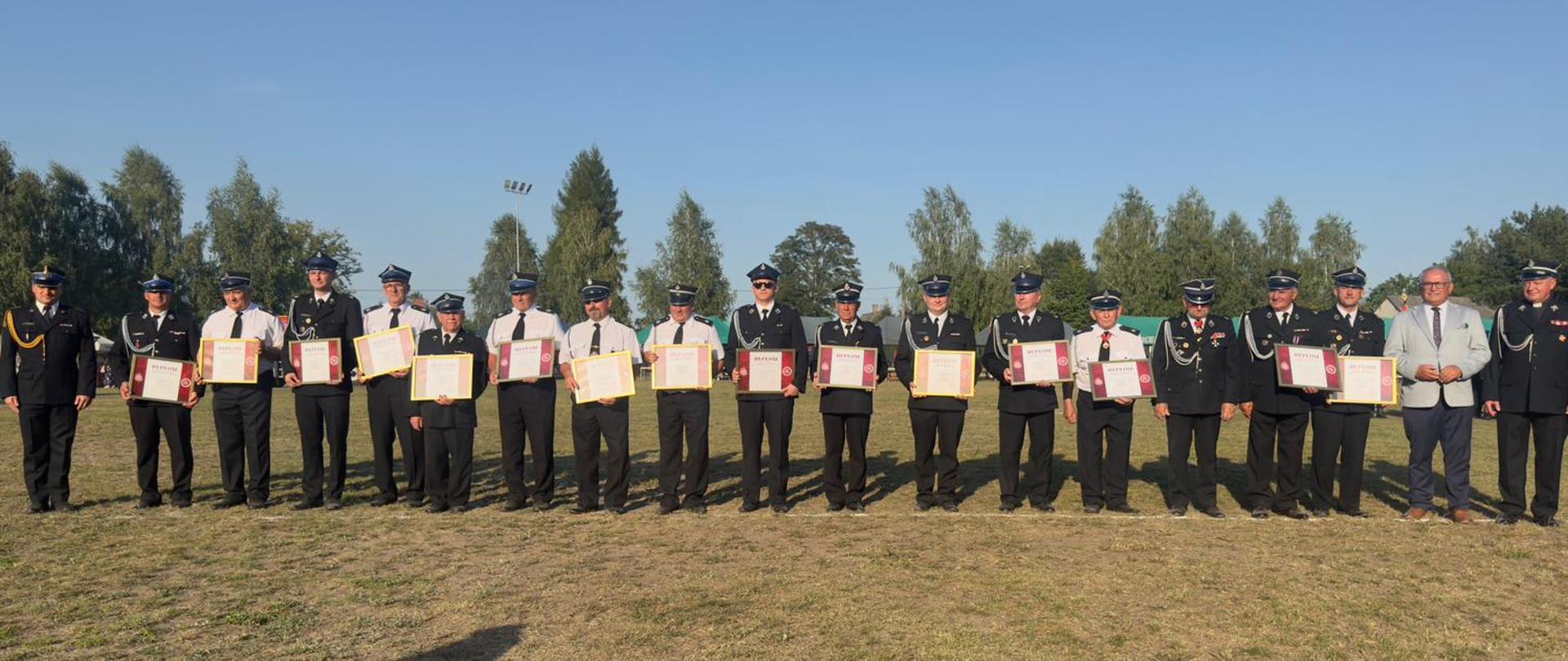 Obchody uroczystości 100-lecia Ochotniczej Straży Pożarnej w Poświętnym. Na pierwszym planie fotografii znajdują się przedstawiciele jednostek Ochotniczych Straży Pożarnych będący w Krajowym Systemie Ratowniczo-Gaśniczym. Zostali oni uhonorowani przez starszego kapitana Krzysztofa Juszczaka Komendanta Powiatowego Państwowej Straży Pożarnej w Opocznie pamiątkowymi dyplomami oraz medalami z okazji 30-lecia Krajowego Systemu Ratowniczo-Gaśniczego. Wszyscy zwróceni są przodem do aparatu, stoją na trawie, ramię w ramię w lekkim rozluźnieniu, ale w wyraźnym szyku. W oddali widoczne są wysokie drzewa, niebo jest bezchmurne, a światło słoneczne tworzy ciepłe, jasne barwy na całym zdjęciu.