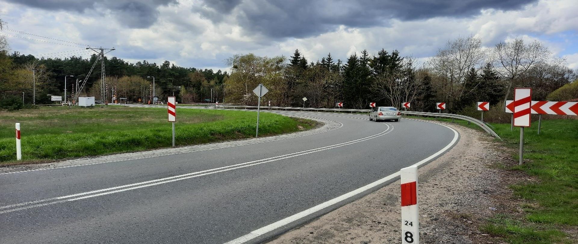 Droga prowadzi ostrym łukiem w kierunku przejazdu kolejowego. Wokół łąki i tereny zadrzewione.