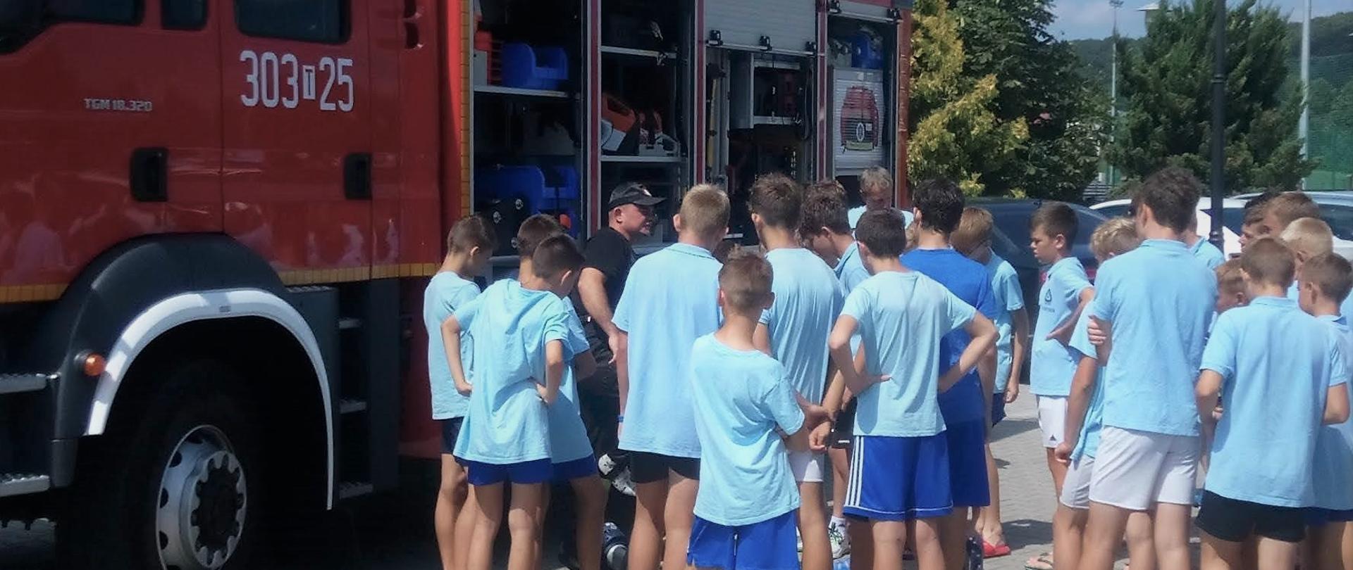 Zdjęcie przedstawia grupę dzieci i młodzieży szkolnej podczas zajęć edukacyjnych ze strażakami przy samochodzie pożarniczym.