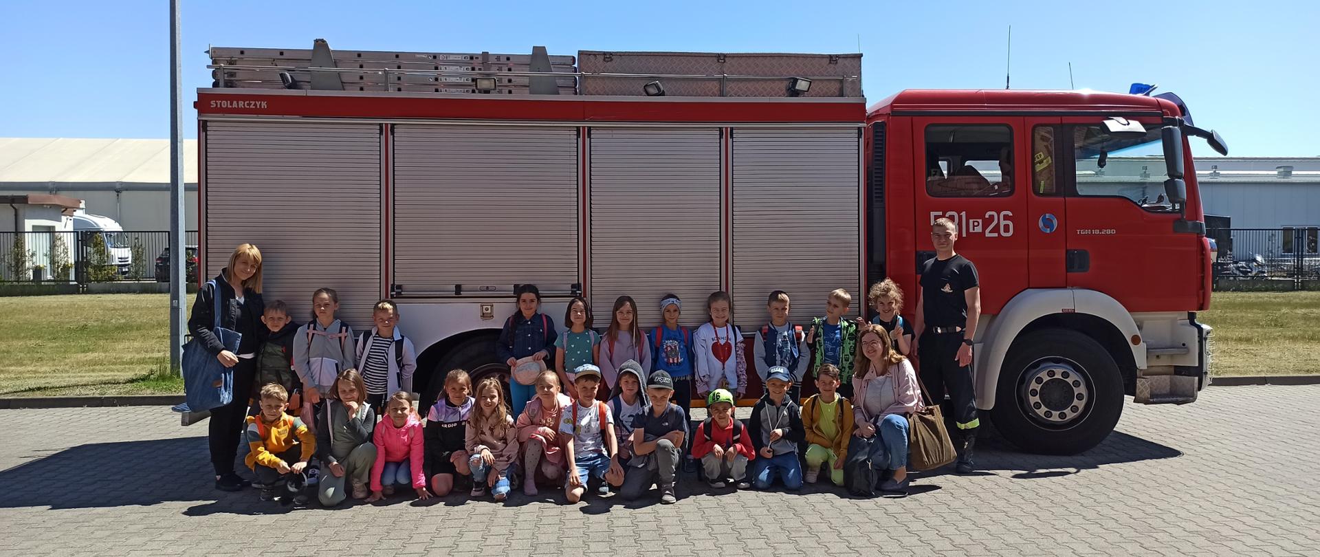 Dzieci, opiekunowie i funkcjonariusz na tle samochodu pożarniczego.